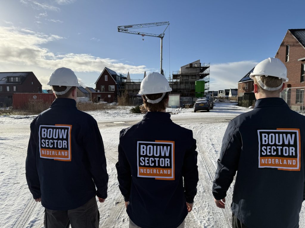 Drie vakmensen van Bouwsector Nederland met witte helmen kijken over een besneeuwde nieuwbouwwijk met een bouwkraan
